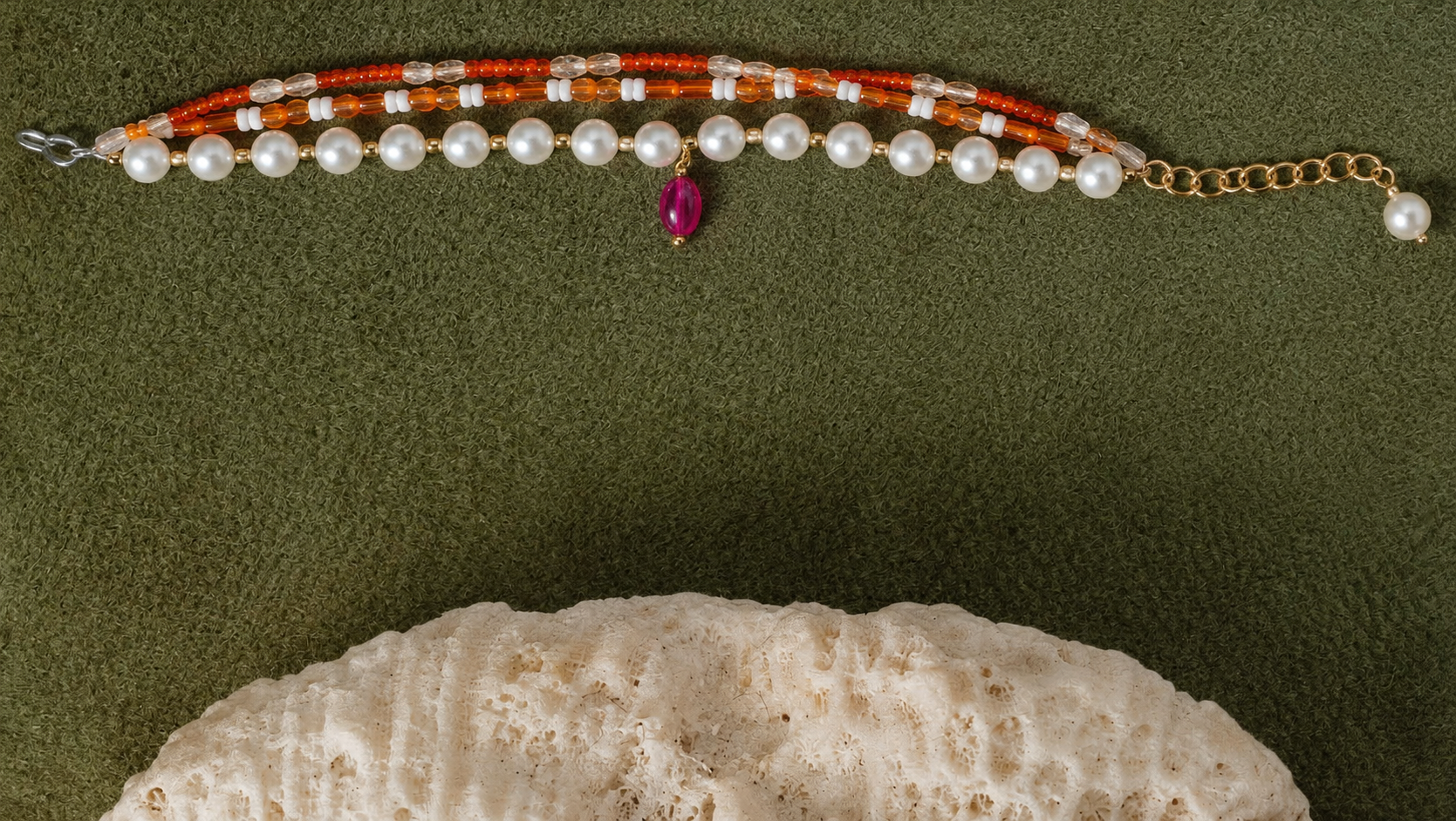 A beaded bracelet with white pearls, orange-red beads, a small pink teardrop charm, and gold-tone chain hardware, laid flat on an olive green background with a white shell prop at the bottom.