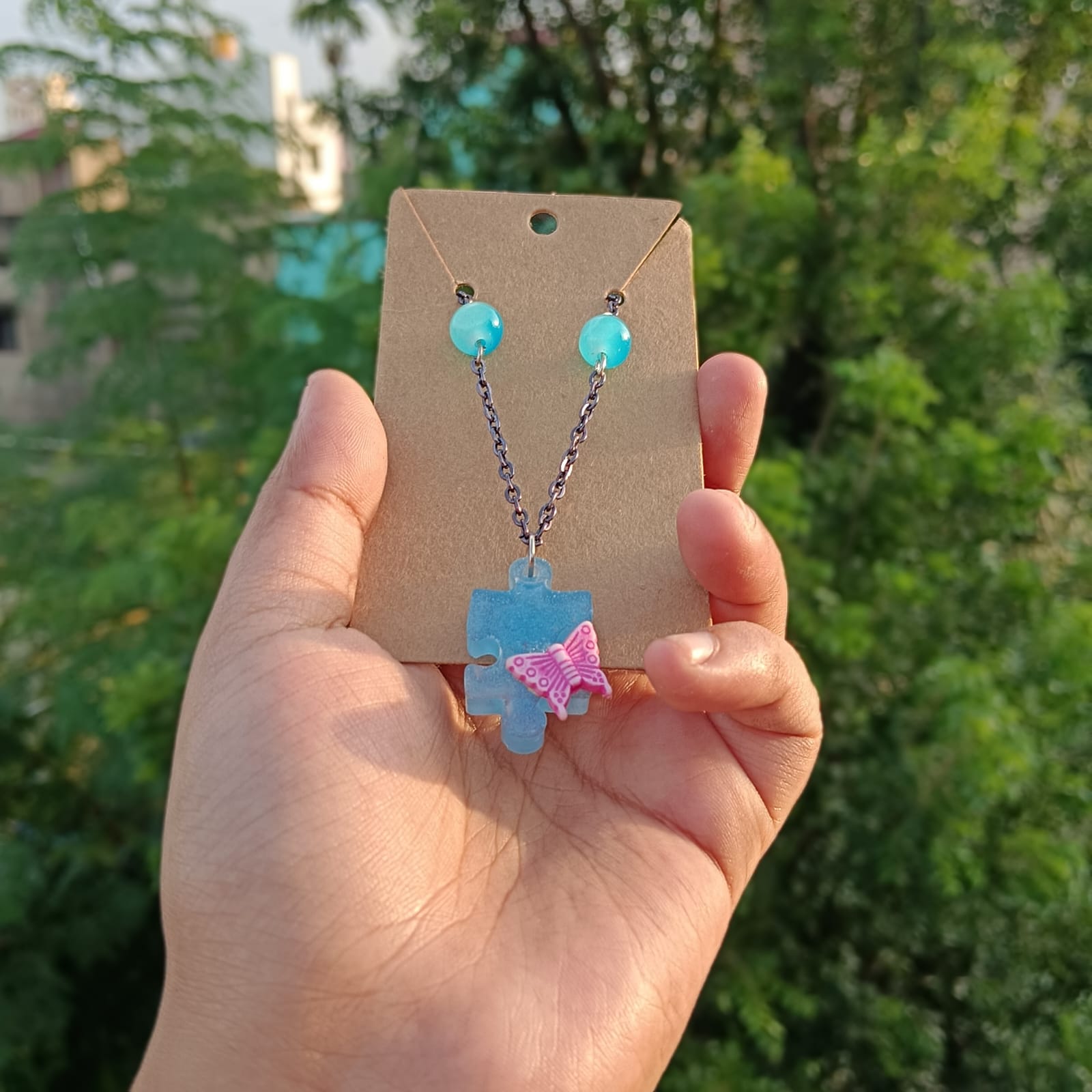 A hand holding a brown display card featuring a light blue puzzle-piece pendant necklace with a pink ribbon accent and matching light blue bead earrings, set against a blurred green outdoor background.