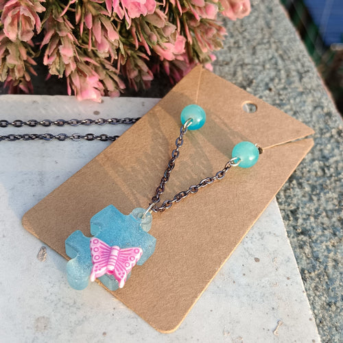 A Handmade resin necklace displayed on a brown kraft card, featuring a translucent light blue puzzle-piece pendant with a pink butterfly accent and light blue bead drop charms on a dark metal chain.