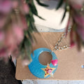 A handmade crescent moon earring with a star and heart charm, displayed on a brown kraft card with blurred pink flowers in the foreground.