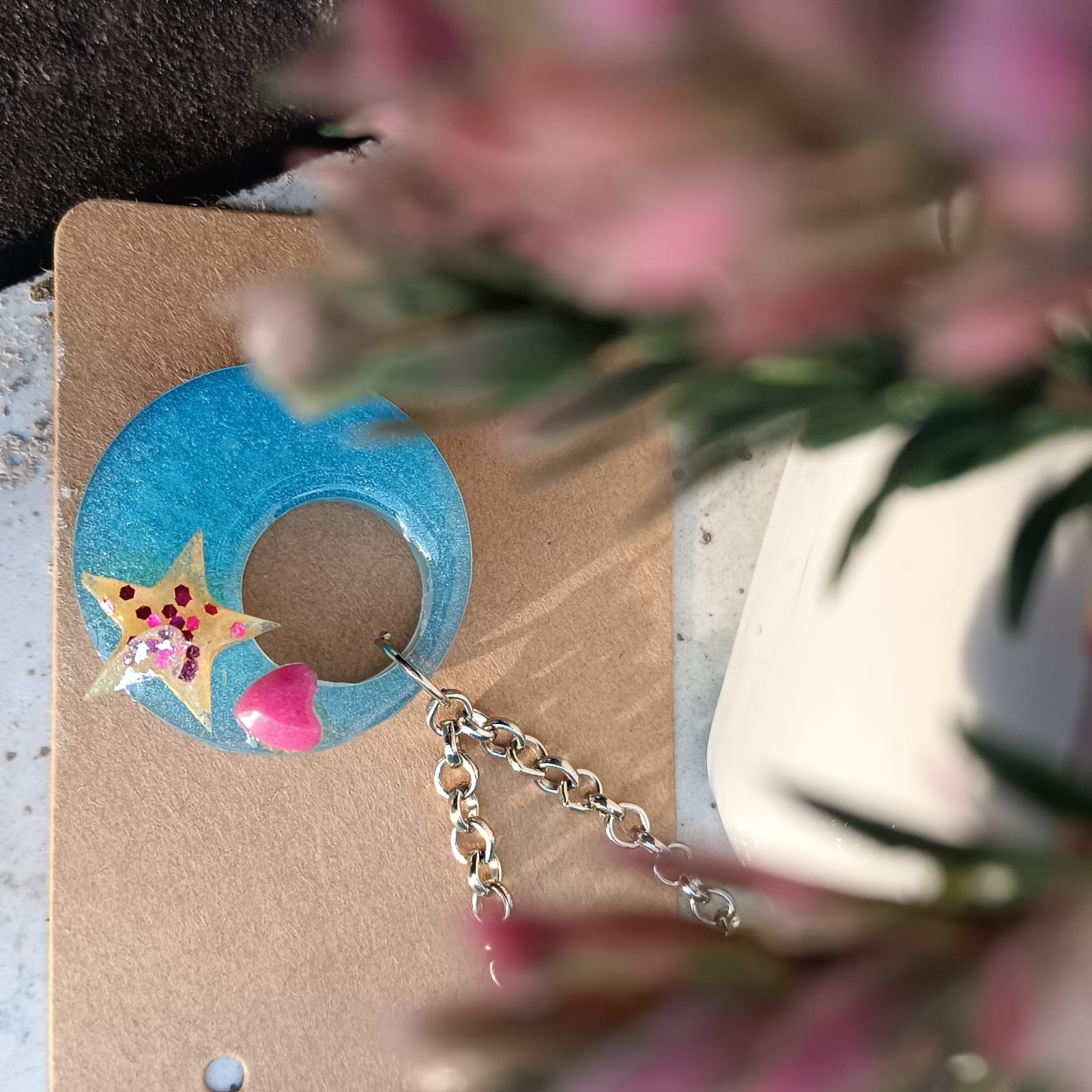 A handmade crescent moon earring with a star and heart charm, displayed on a brown kraft card with blurred pink flowers in the foreground.