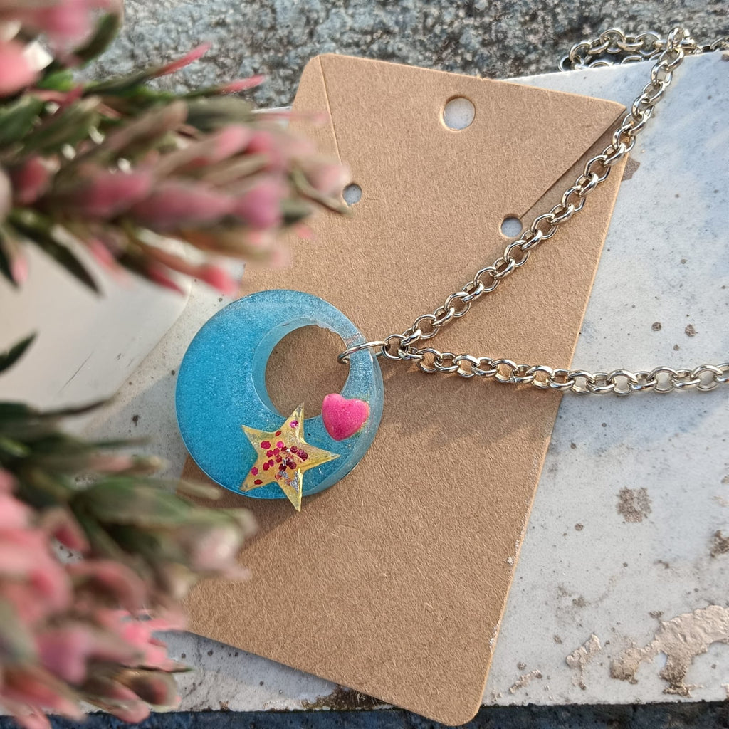 A necklace with a light blue crescent moon pendant featuring a yellow star and a pink heart, displayed on a kraft paper card with blurred pink flowers in the foreground.